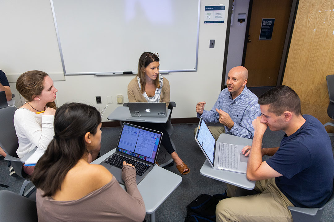 Professor Steven Balla and crouches down in a classroom to talk to a group of students seated in a circle of desks