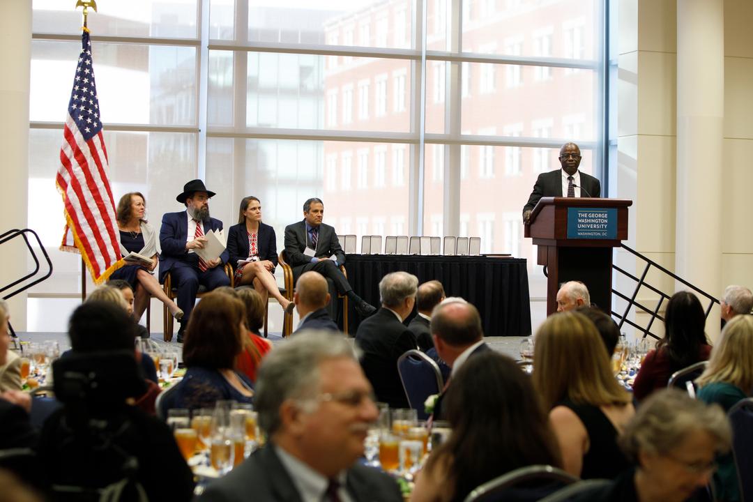 A man in a suit stands at a podium on stage addressing a crowd, with banquet tables and American flags decorating the room