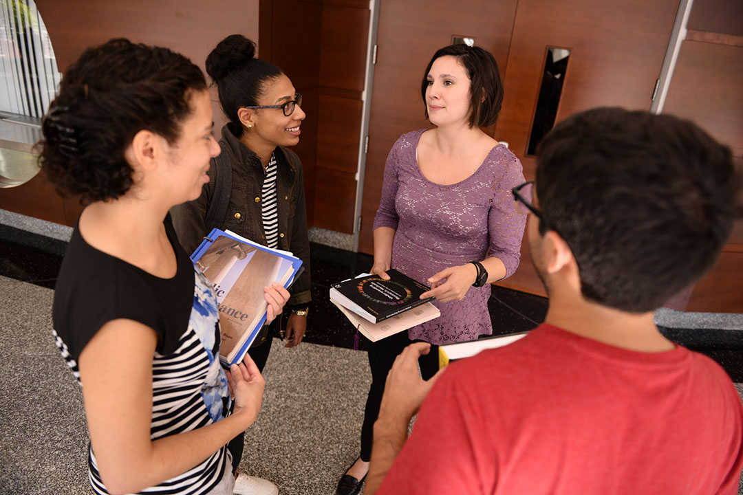A group of four students viewed from above chatting in a hallway outside a classroom holding books