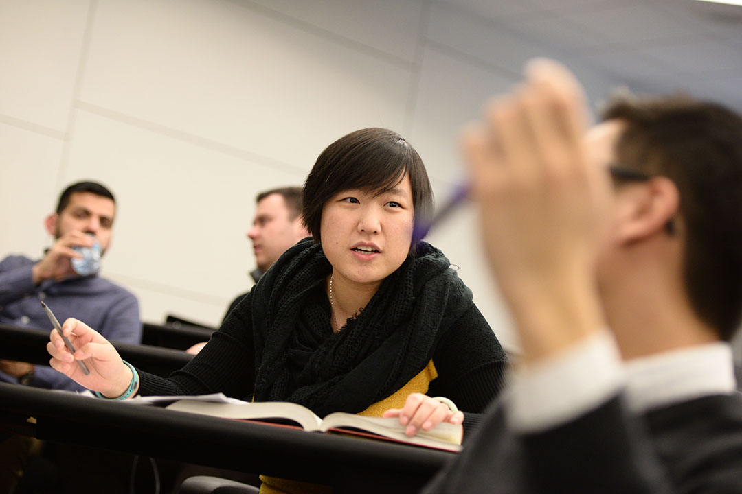 Image of a student in class talking and holding a pencil while her classmates listen