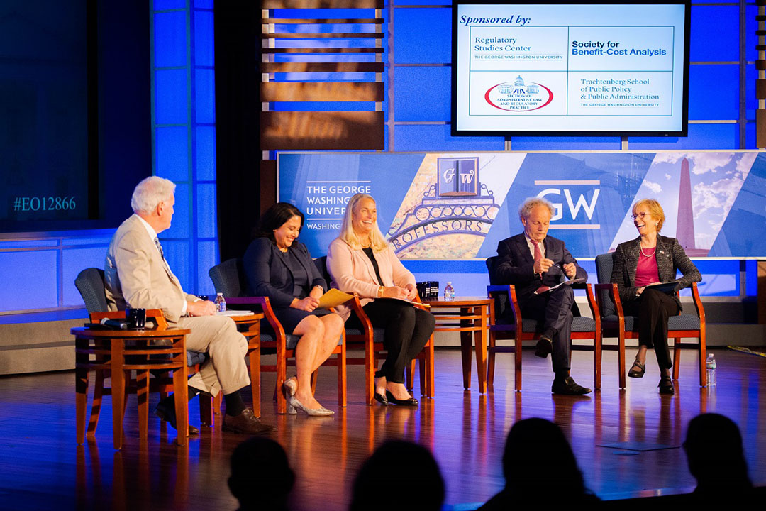 A group of speakers on stage in chairs talking with GW and Regulatory Studies Center signage in the background