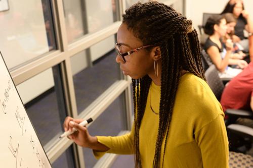 A woman in braids and a yellow sweater writes on a whiteboard in a small classroom
