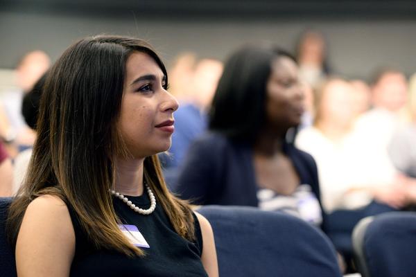 A woman in profile seated in an auditorium wearing professional clothing and a name tag