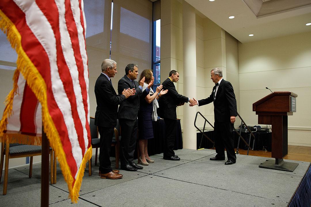 Three TSPPPA faculty members on stage next to an American flag clapping and presenting an award to an awardee