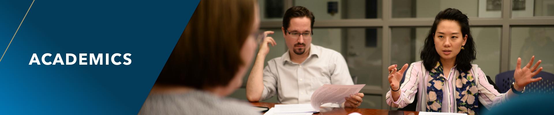 Academics. An Asian American woman gesturing and talking at a shared table, while students around her listen.