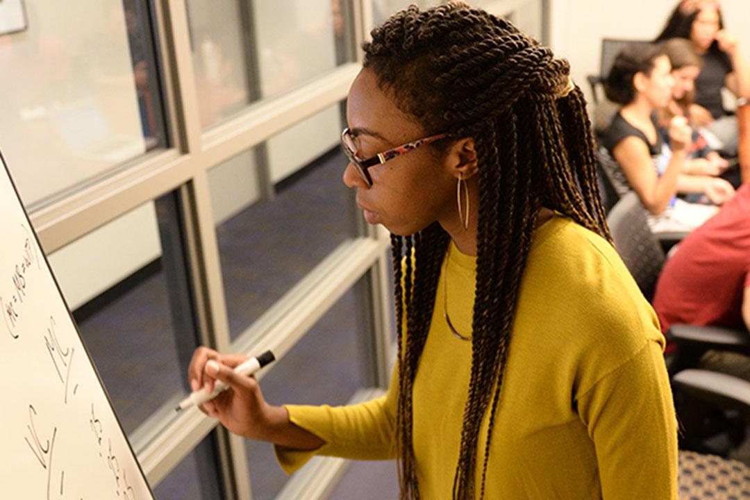 A woman in braids and a yellow sweater writes on a whiteboard in a small classroom