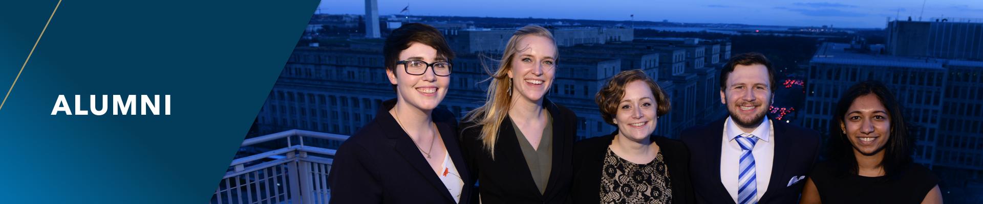 Alumni. A group of five students in business casual dress stand on the rooftop of a GW building at night with the DC skyline in the background